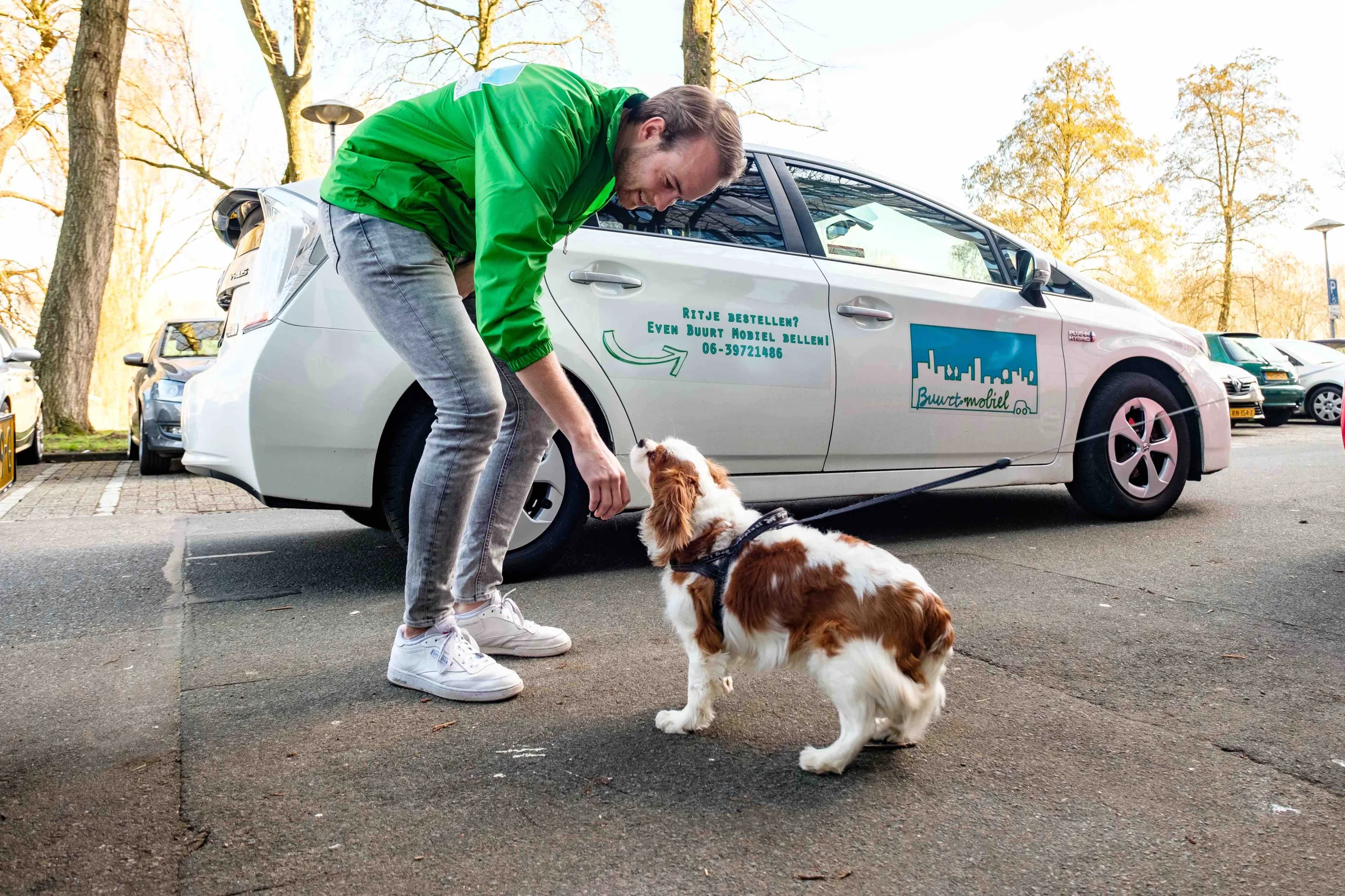 Chauffeur van gemeenschapsvervoerservice Buurt Mobiel aait hond voor witte auto van Buurt Mobiel