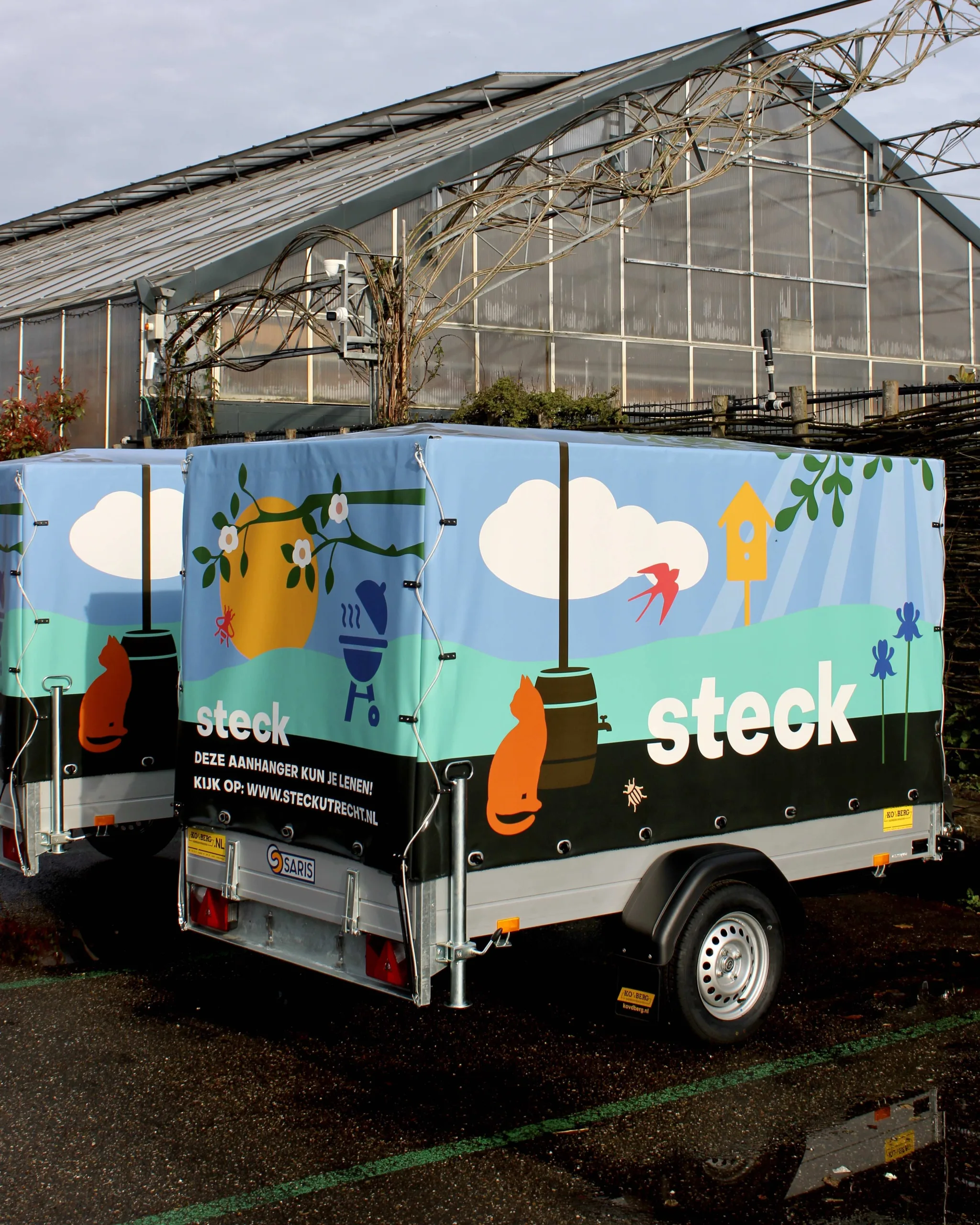 Aanhangwagen van tuincentrum Steck staat op het parkeerterrein voor de kas. De aanhangwagen heeft een kleurrijke en zomers gedecoreerde buitenkant, waar ook het logo van Steck op staat