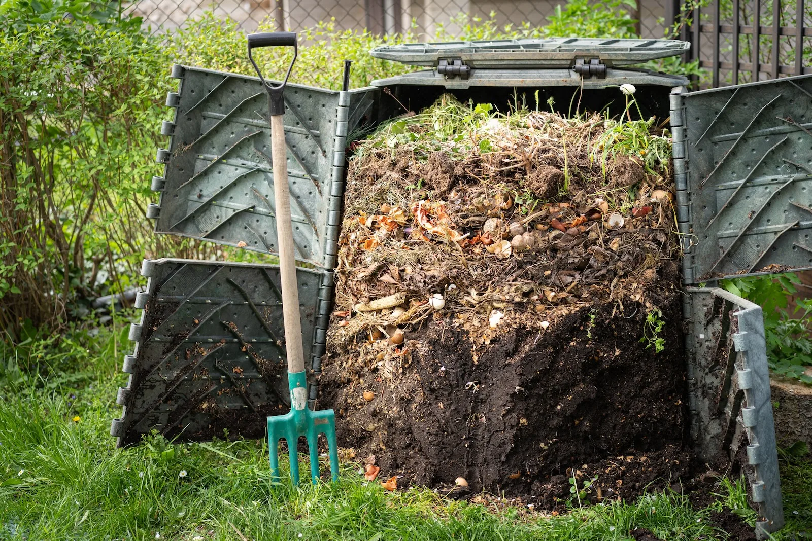 Een grote compostbak van donkergroene kleur, gevuld met lagen organisch afval, staat in een achtertuin. Het materiaal in de bak bevat een mix van aarde, plantenresten, en keukenafval zoals groenteschillen. Een deel van de compost is al afgebroken tot donkere, rijke aarde. Naast de compostbak staat een tuinvork met een houten steel en groene tanden, waarschijnlijk gebruikt om het compostmateriaal om te keren. De bak is omgeven door een weelderige groene tuin en een hek op de achtergrond, wat suggereert dat dit een actief gebruikt en goed onderhouden compostsysteem is in een huiselijke omgeving.