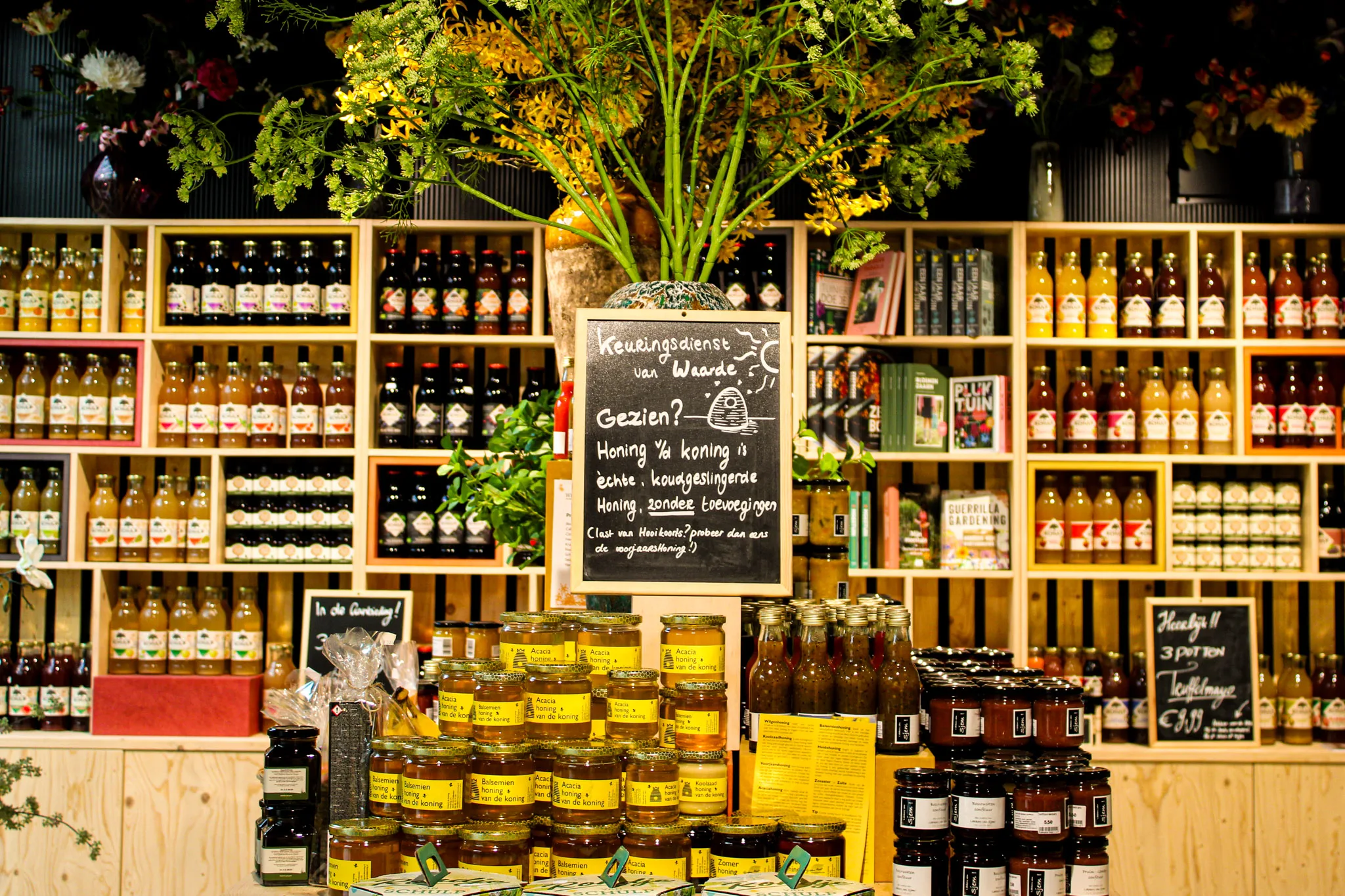 Een netjes gerangschikte houten schap met talloze flessen Schulp sap en potten verse honing, in de winkel van Oogst in Steck Utrecht. Middenin staat een krijtbord met tekst over de kwaliteit van honing.