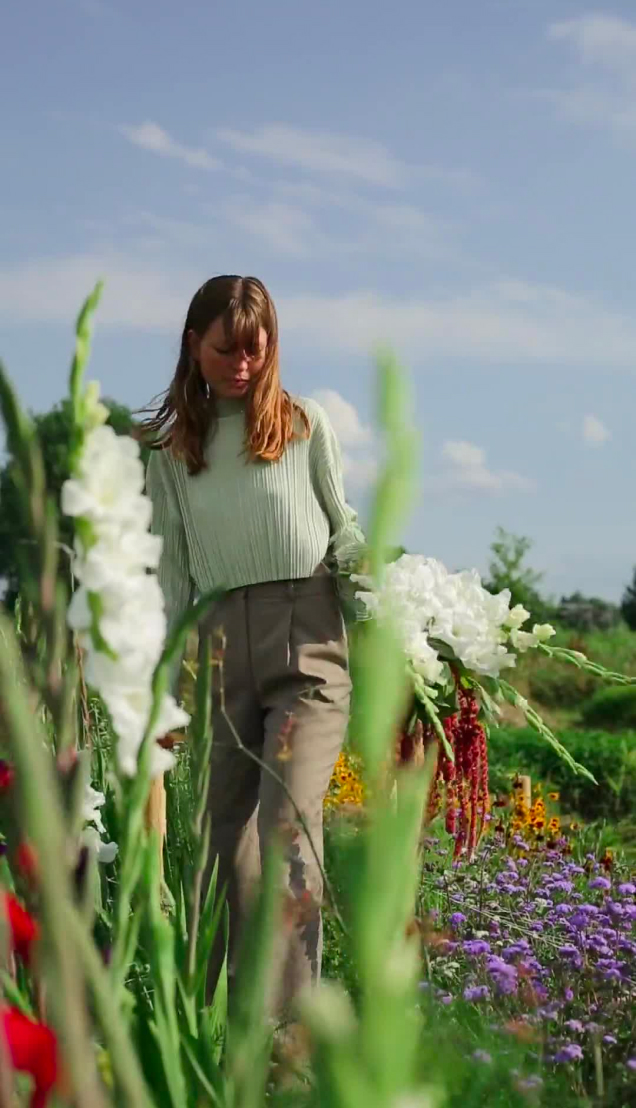 Noraly Rozendaal loopt door de bloemenpluktuin met een grote bos witte bloemen in haar handen. Ze draagt een lichtgroene trui en beige broek. De tuin om haar heen staat vol met kleurrijke bloemen, waaronder paarse en rode bloemen, onder een heldere blauwe hemel.