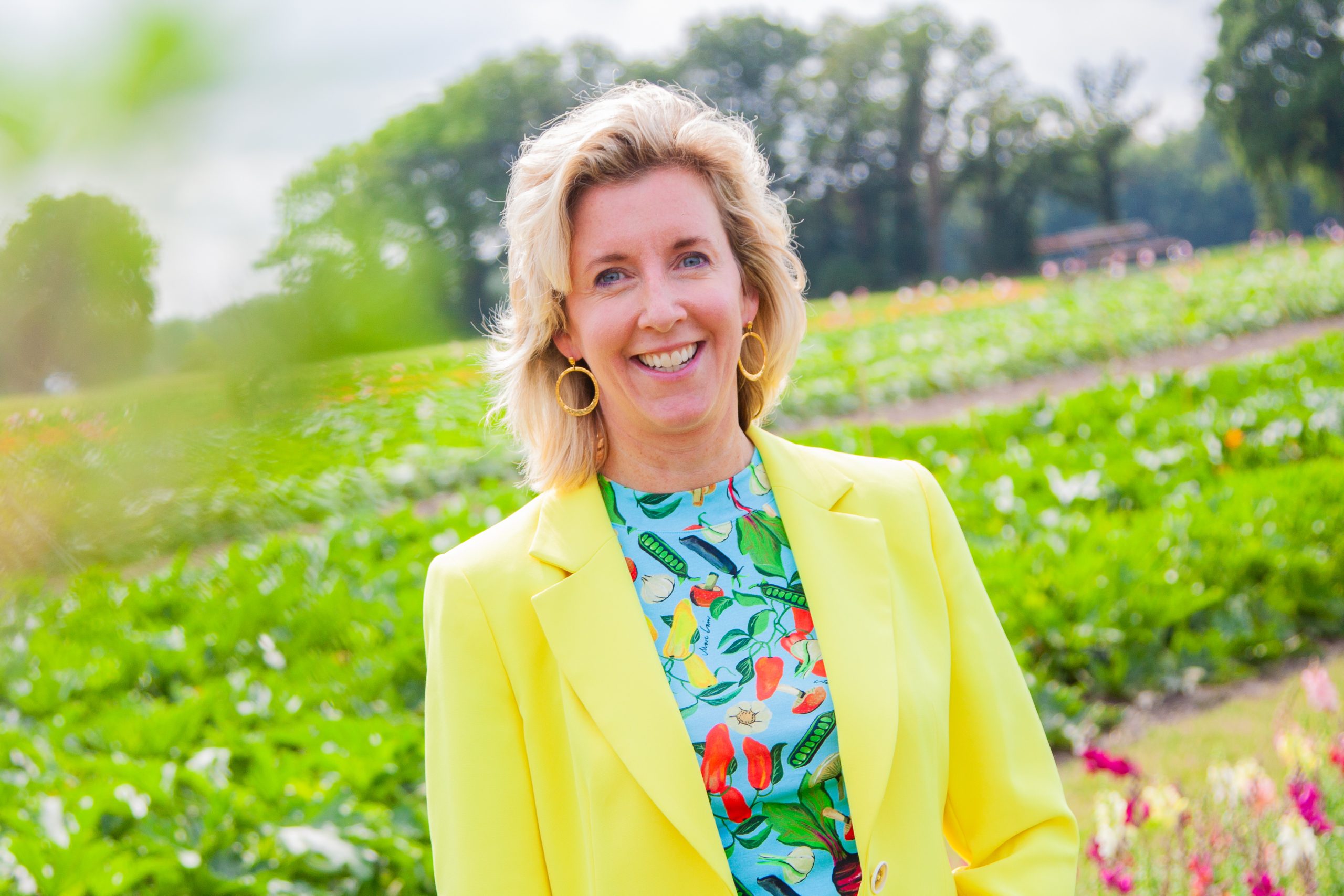 Een vrolijke vrouw met blond haar staat glimlachend voor een groene achtergrond van een bloeiend veld. Ze draagt een felgeel colbert en een kleurrijke blouse met een patroon van verschillende groenten en vruchten. Op de achtergrond zijn bomen en een heldere lucht zichtbaar, wat de setting een zomers en natuurlijk gevoel geeft.