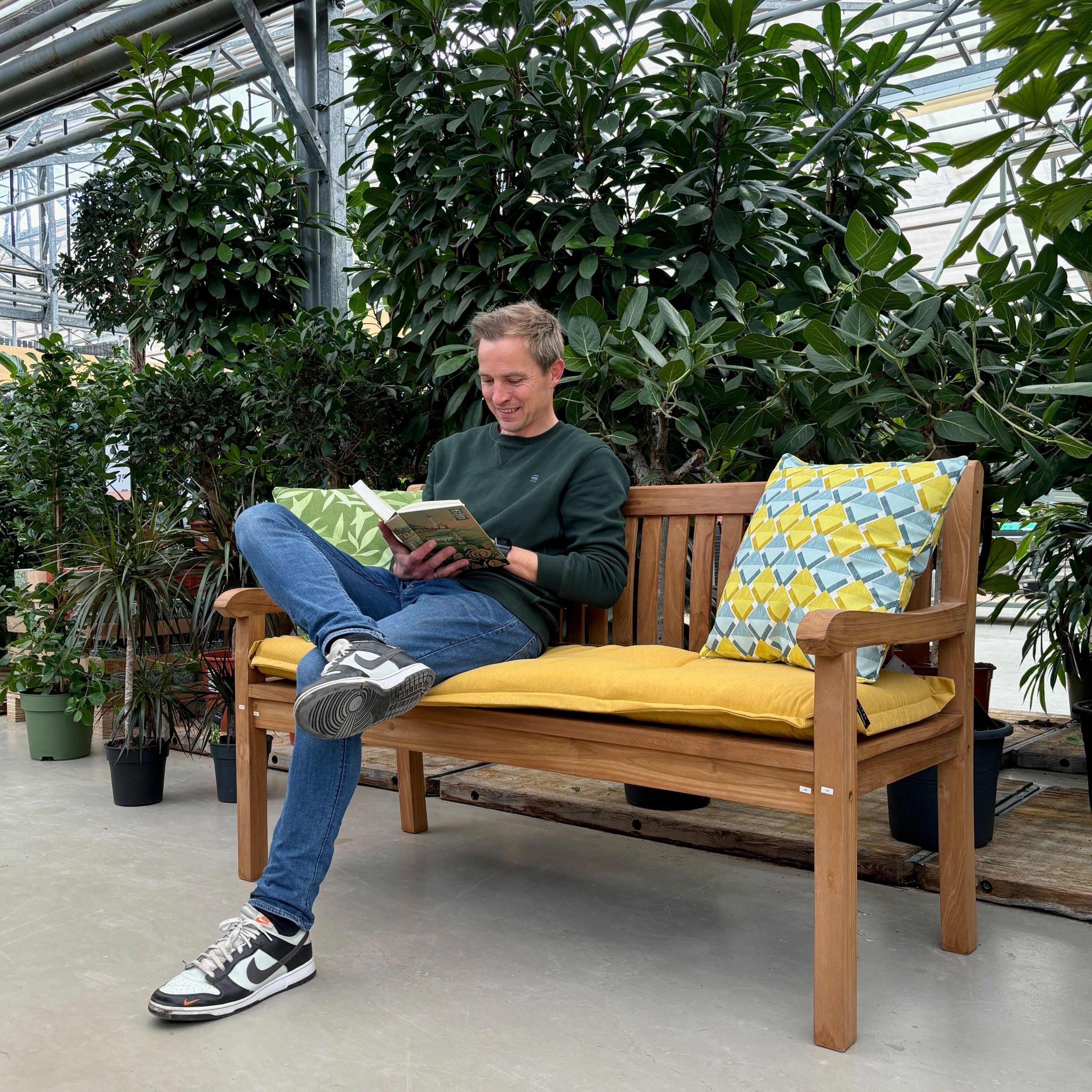 Een man zit ontspannen op een houten tuinbank met gele kussens in de tuinmeubelenafdeling van Steck in Utrecht Overvecht. Hij leest een boek en lijkt te genieten van het moment. De bank is omringd door groene planten en potten, die zorgen voor een natuurlijke en rustgevende sfeer. De serreachtige omgeving met glazen dak en stalen constructies is ook zichtbaar. De man draagt een donkere trui, blauwe jeans en sneakers.