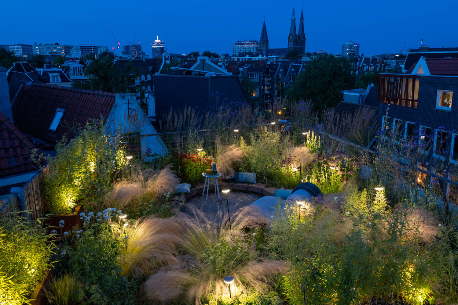 Een sfeervol verlicht groendak in de avond, omringd door oude stadspanden. Het dak is gevuld met diverse siergrassen en planten, en er zijn enkele zitplekken gecreëerd. De verlichting zorgt voor een warme, uitnodigende sfeer.