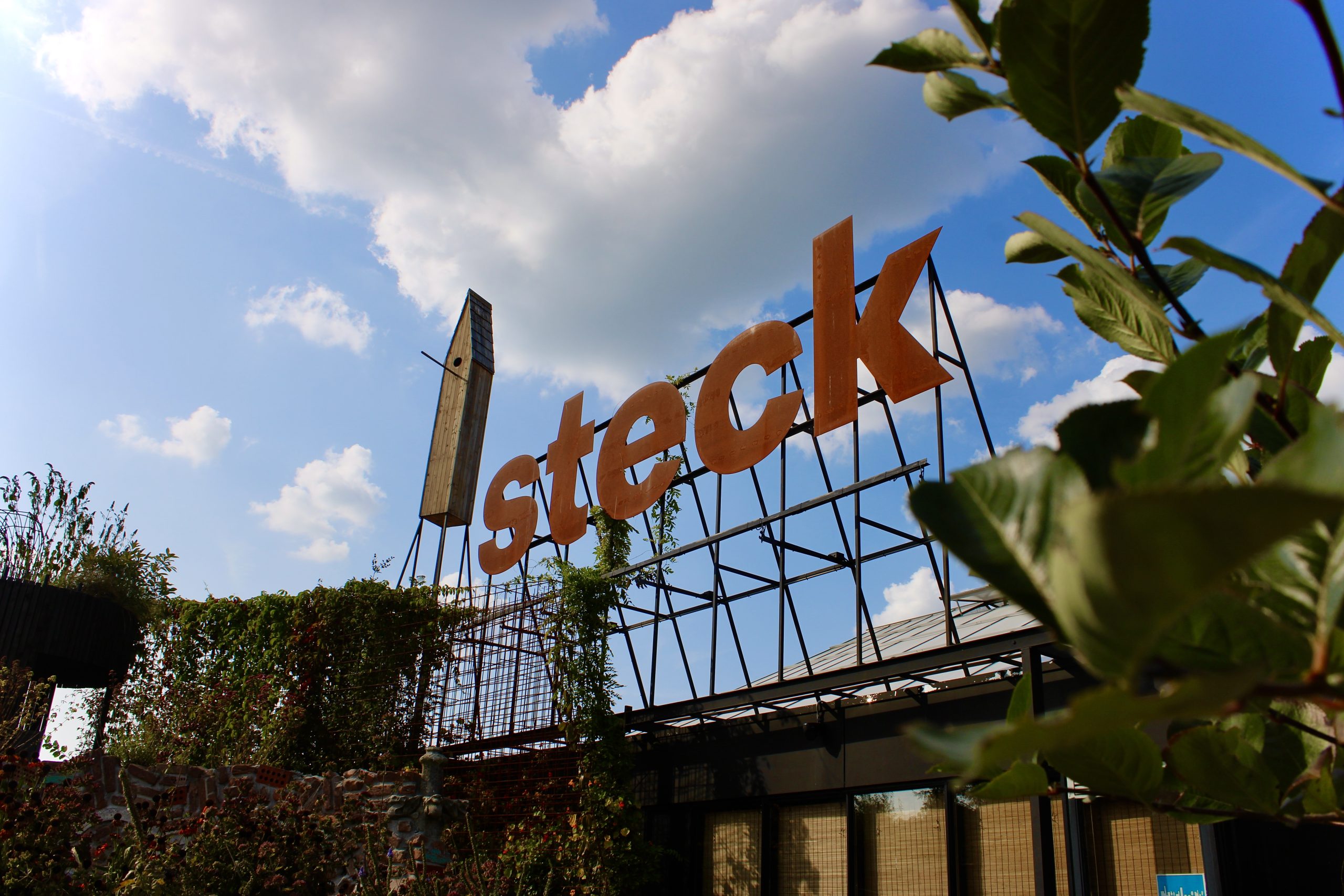 Hoofdingang van tuincentrum Steck in Utrecht Overvecht in zomerse omstandigheden. Rond de grote letters met de naam groeien planten, daarachter een blauwe lucht met een paar wolkjes. Het karakteristieke grote vogelhuis staat naast de letters