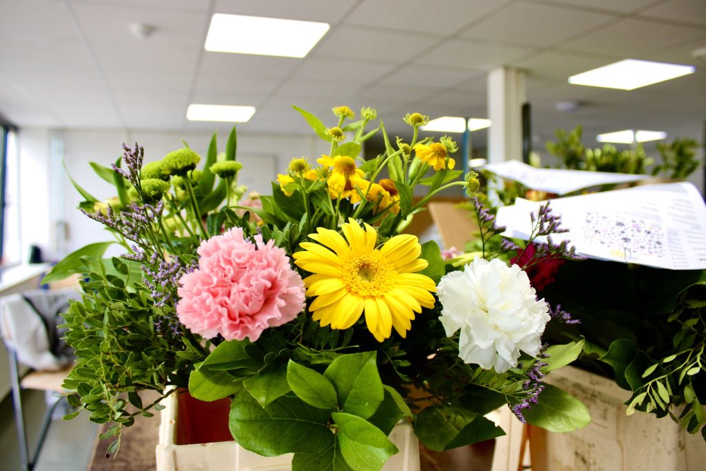 Een kleurrijk, vers samengesteld boeket met onder andere roze, gele en witte bloemen. Het boeket staat op een tafel in een werkruimte.