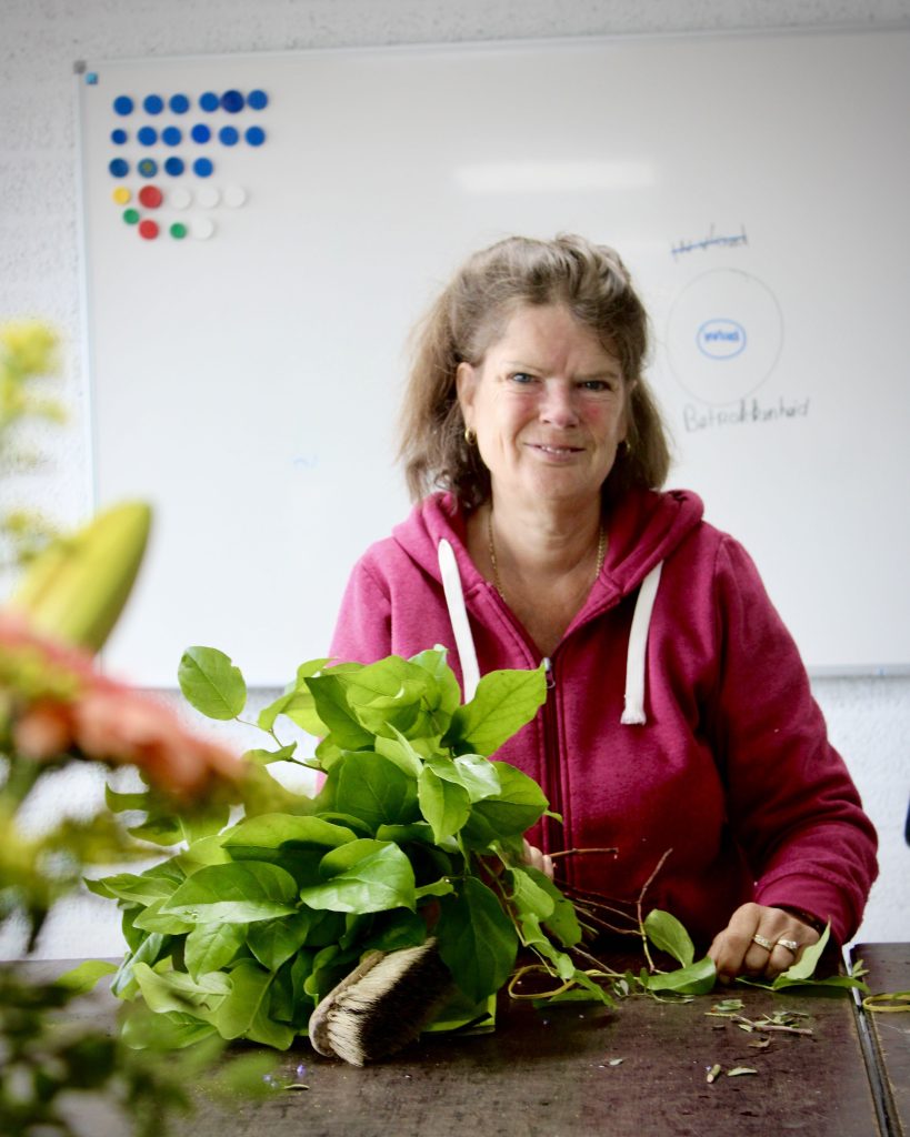 Een medewerker van UW Flora zit aan een tafel met groene takken en bladeren, klaar om een boeket samen te stellen. Ze draagt een roze trui en glimlacht vriendelijk naar de camera. Op de achtergrond staat een wit schoolbord met magneten.