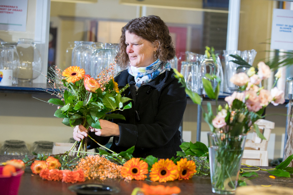 Een bloemiste, gekleed in een zwarte jas en een blauwe sjaal, is geconcentreerd bezig met het samenstellen van een kleurrijk boeket. Op de tafel voor haar liggen verschillende bloemen, waaronder oranje gerbera's, klaar om verwerkt te worden in het boeket. De achtergrond toont een werkruimte met glazen vazen en bloemgereedschap, wat de professionele omgeving van de bloemisterij benadrukt.