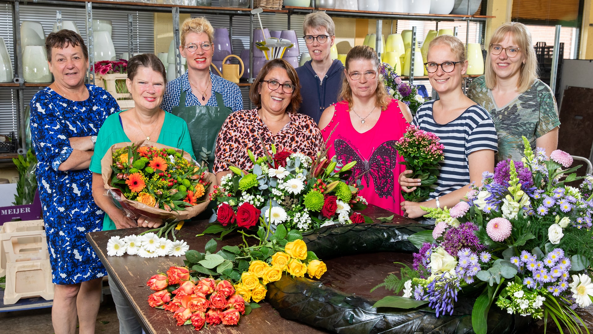 Een groep van acht bloemisten poseert samen achter een tafel vol kleurrijke bloemen en bloemstukken. Ze lachen en stralen trots uit. Voor hen liggen diverse bloemen, waaronder rozen, gerbera's, en chrysanten, op de tafel. De ruimte waarin ze staan is gevuld met vazen en bloemgerelateerde materialen. De sfeer is warm en gezellig, wat de samenwerking en het vakmanschap van de groep benadrukt.