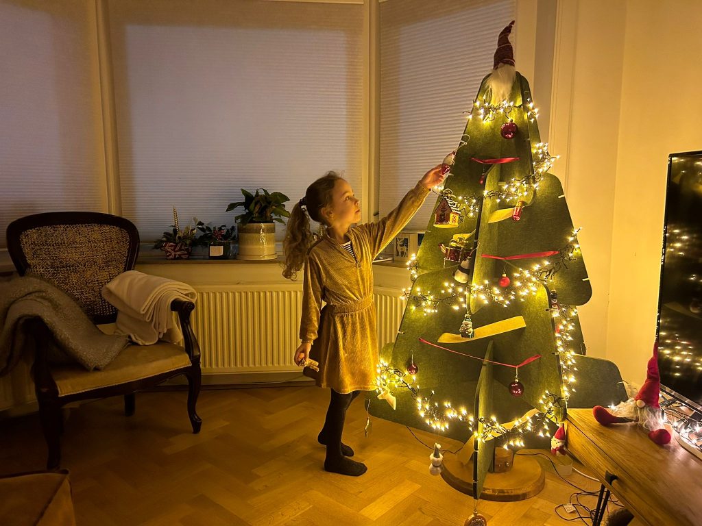 Een meisje in een feestelijke jurk bewondert een verlichte, groene vilten kerstboom van Jingle Tree in een sfeervol verlichte woonkamer. De boom is versierd met lichtjes en kleine ornamenten, en de warme gloed van de lampjes geeft een knusse en feestelijke sfeer aan het geheel.