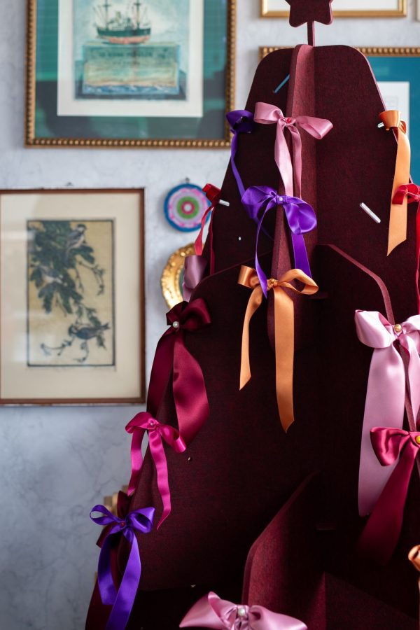 Een close-up van een rode vilten kerstboom van Jingle Tree, versierd met gekleurde satijnen strikken in tinten zoals paars, roze en oranje. De achtergrond toont kunstwerken aan de muur, wat een artistiek en huiselijk gevoel aan de setting toevoegt.