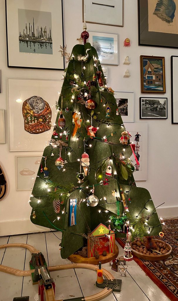 Een groene vilten kerstboom van Jingle Tree staat versierd in een gezellige woonkamer. De boom is gedecoreerd met traditionele kerstballen, kleine figuurtjes en lichtjes die hem een warme sfeer geven. Onder de boom ligt een houten speelgoedtrein en enkele natuurlijke decoraties zoals walnoten.